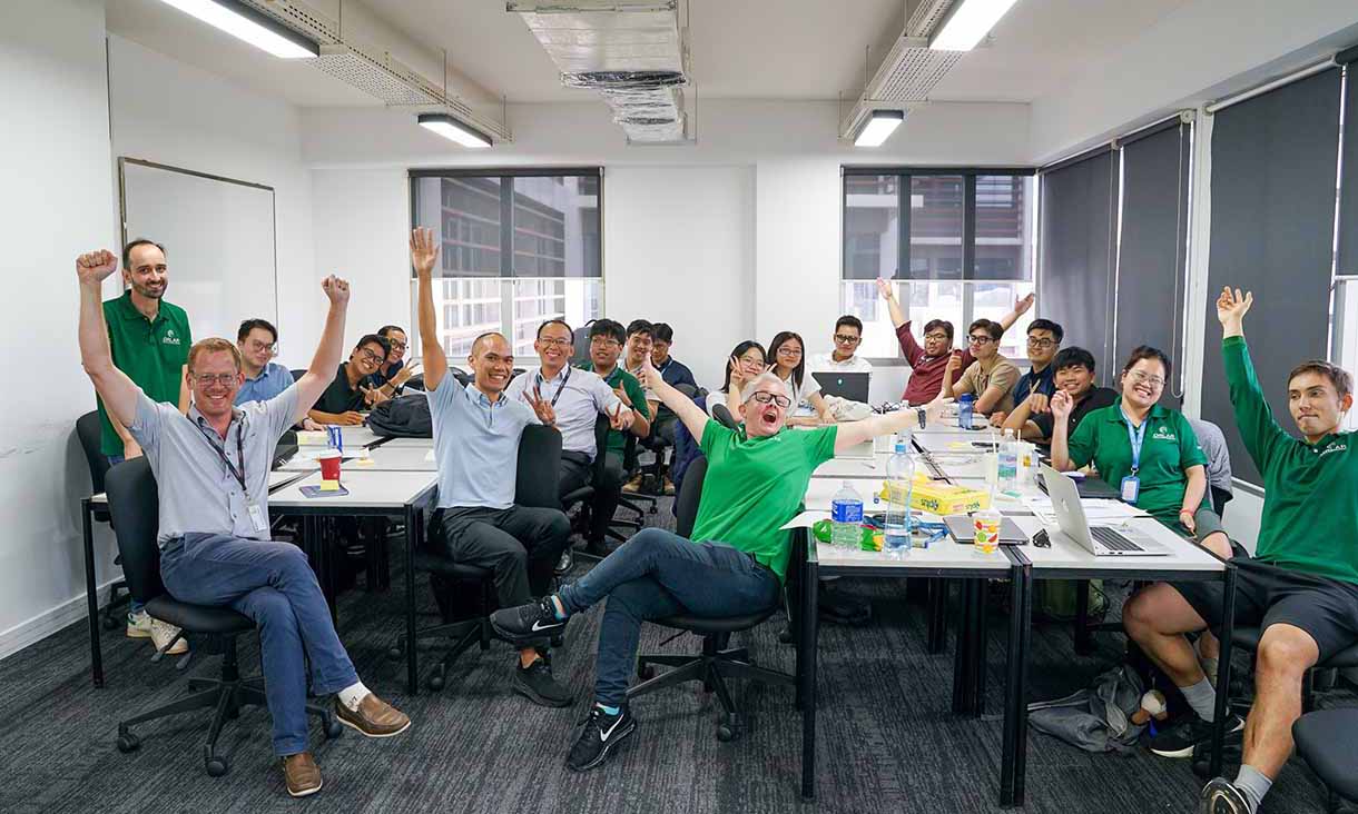 A group of people in a classroom sitting around tables, smiling and raising their arms in celebration. Laptops, drinks, and papers are on the desks, showing a positive and collaborative atmosphere.