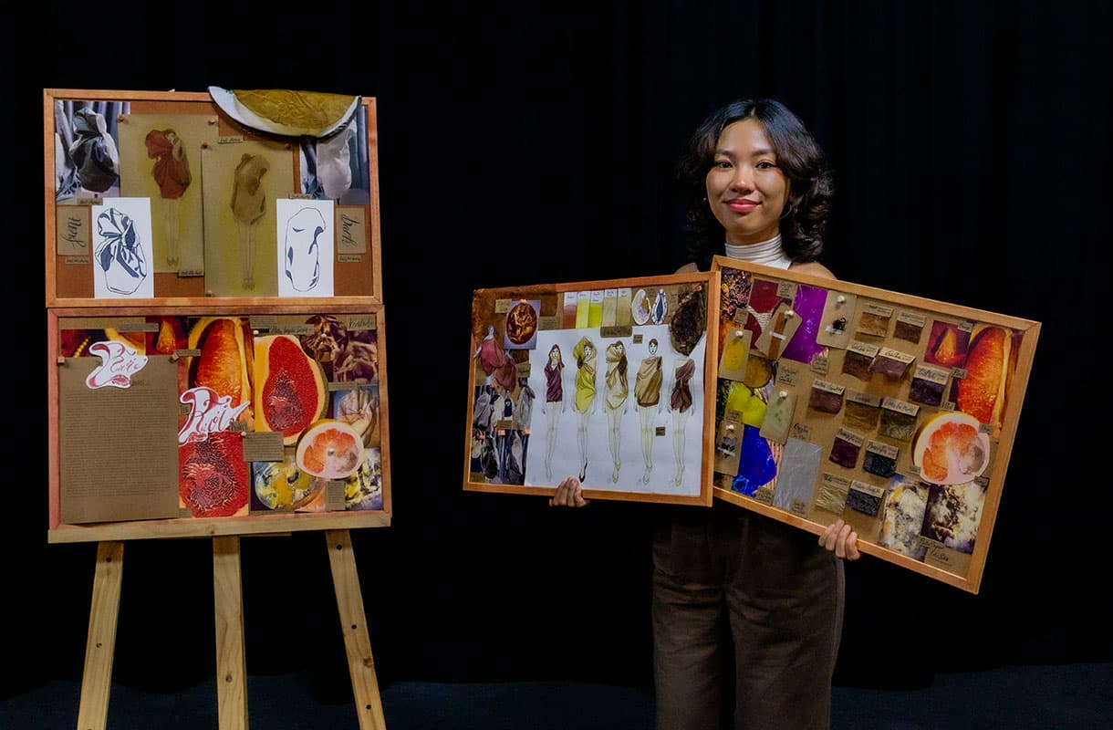 A person stands in front of a black background, holding two boards displaying fashion sketches, fabric samples, and images, with another board on an easel featuring drawings and vibrant fruit imagery.