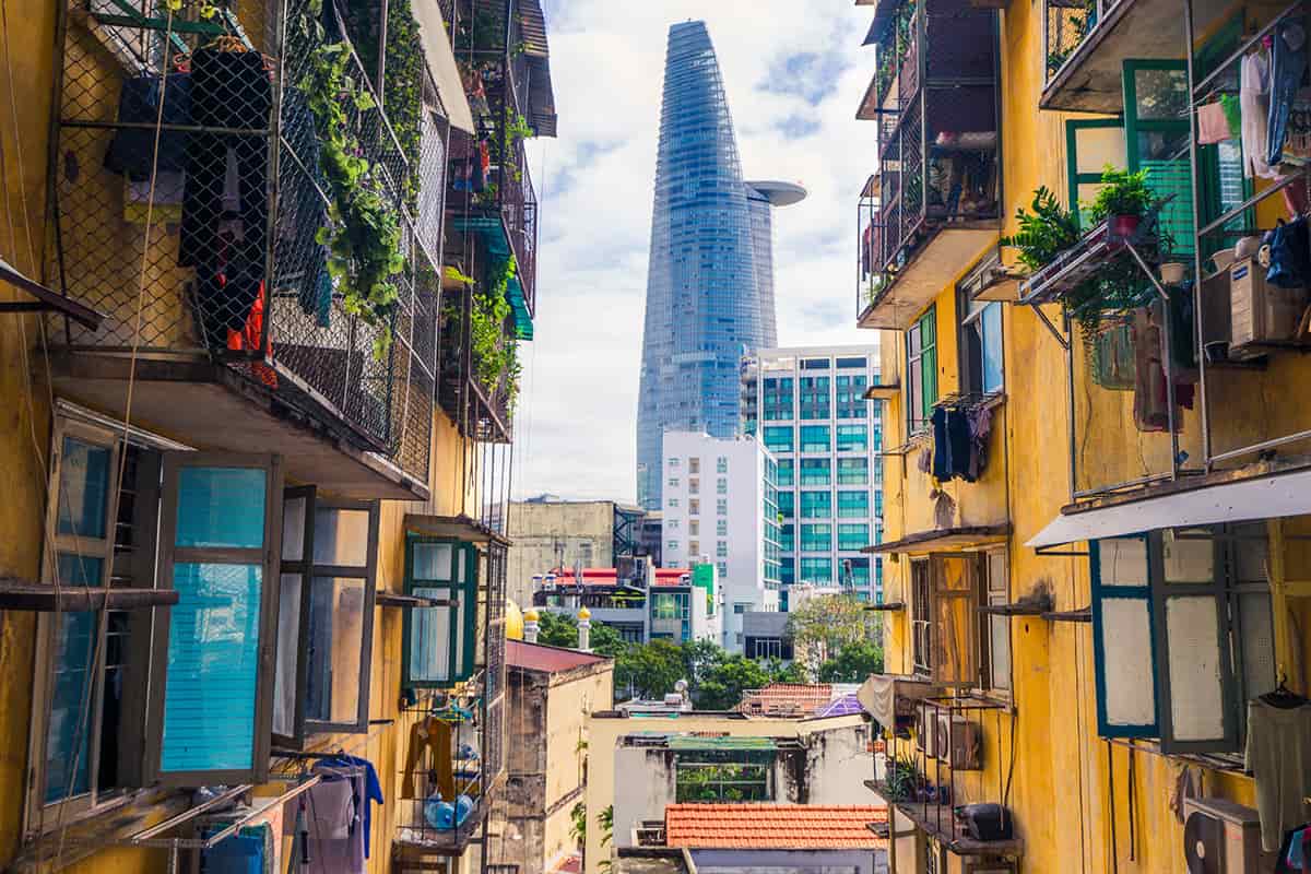 View between two yellow apartment buildings with open windows and balconies, looking toward a modern glass skyscraper in the city centre under a partly cloudy sky.