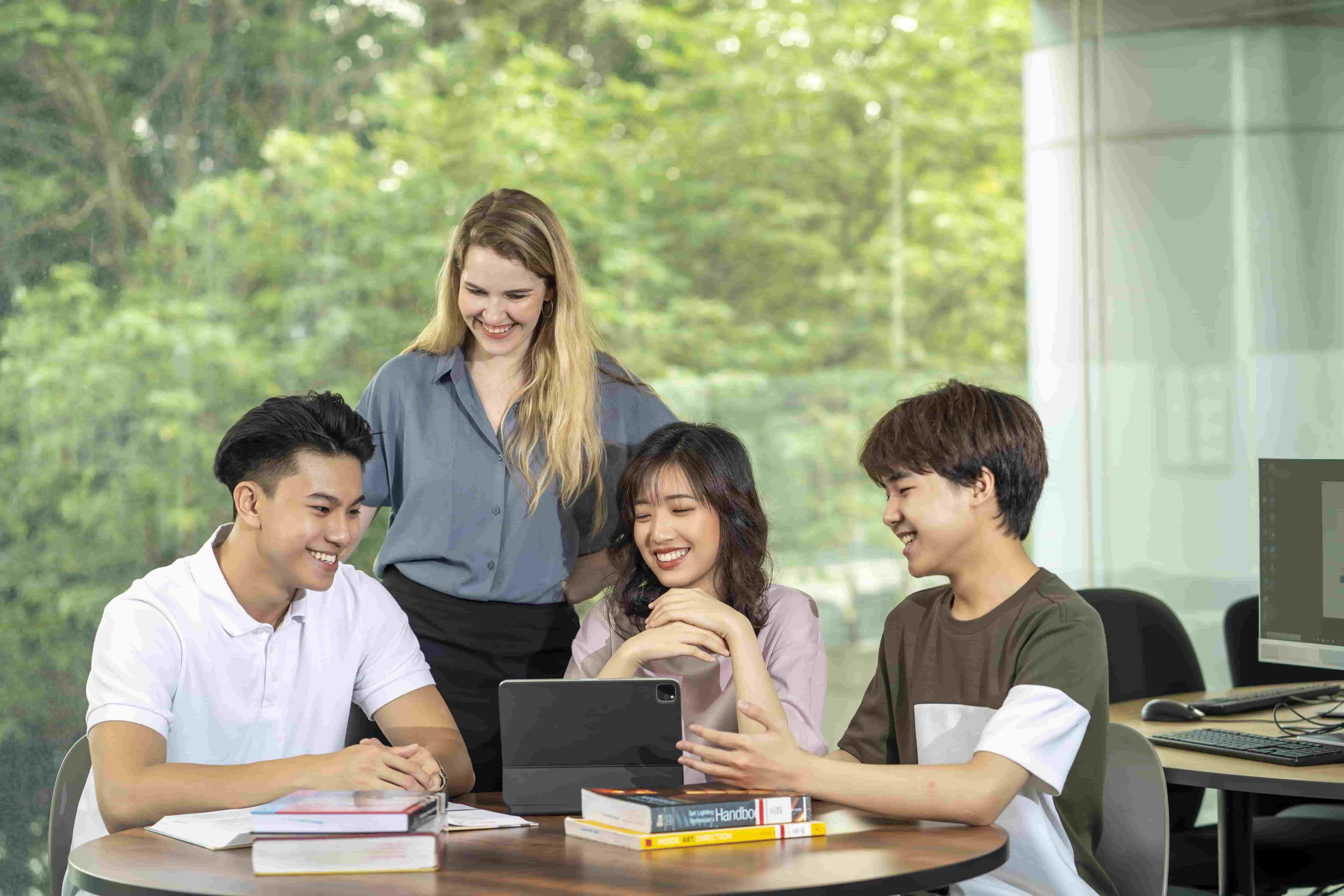 three students sit together on a table and a female student stands next to them and smile