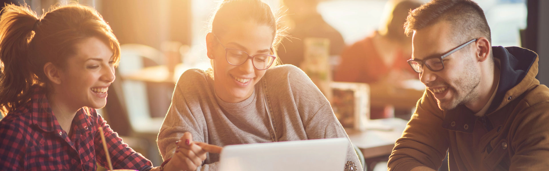 Blonde student wearing glasses and wearing a beige woolen jumper smiling at the laptop, the image is heavily filtered with afternoon sunlight.