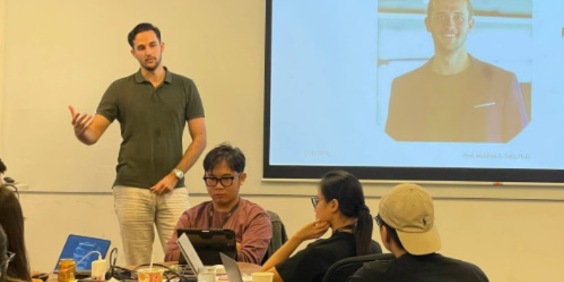 Photo of a guest lecturer presenting in front of the interactive whiteboard to the students who are sitting with their electronic devices