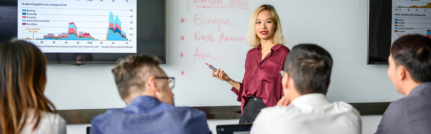 postgraduate group in class room, engaged in a discussion with visual data on a large screen and notes on a whiteboard.