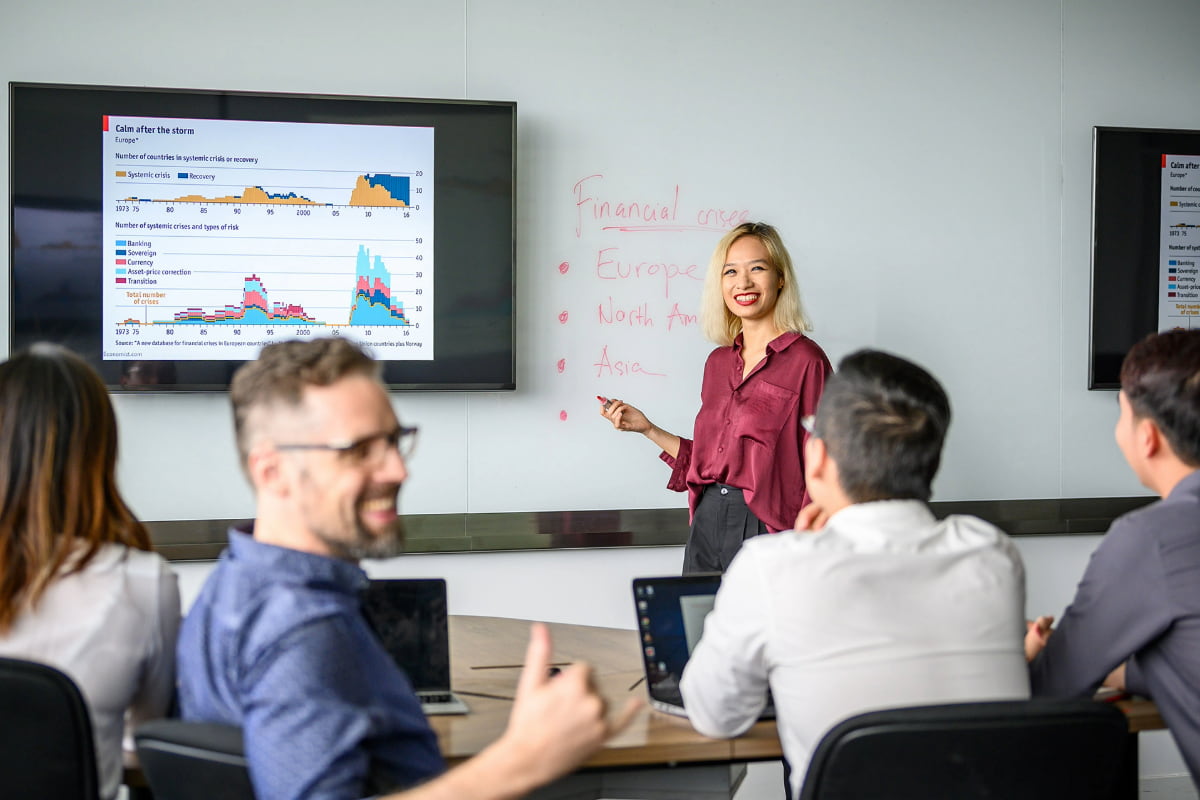 postgraduate group in class room, engaged in a discussion with visual data on a large screen and notes on a whiteboard.
