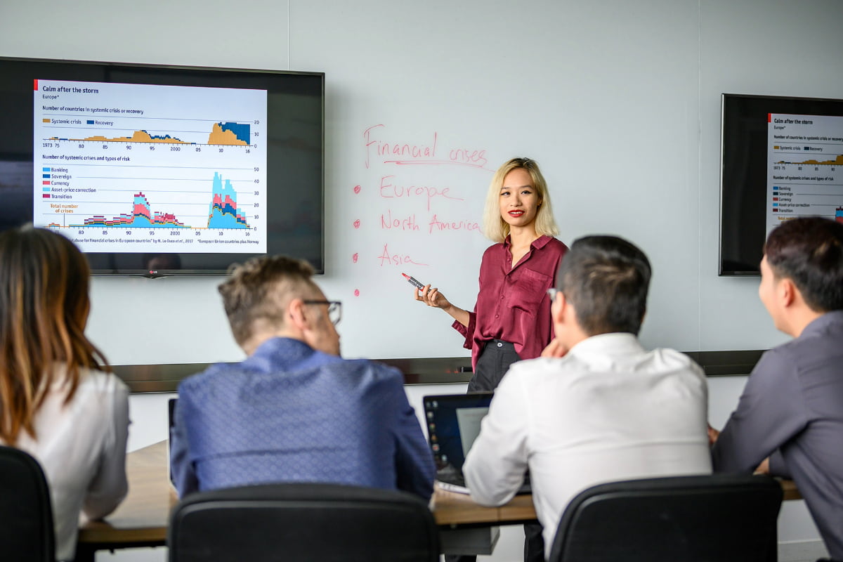 postgraduate group in class room, engaged in a discussion with visual data on a large screen and notes on a whiteboard.