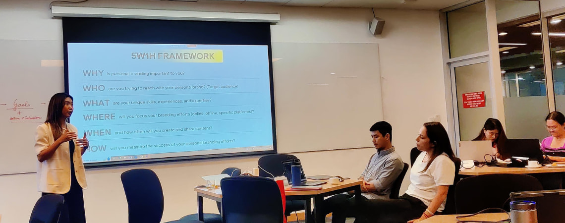 Photo of a guest lecturer presenting in front of the interactive whiteboard titling 5w1h framework to the students who are sitting with their electronic devices
