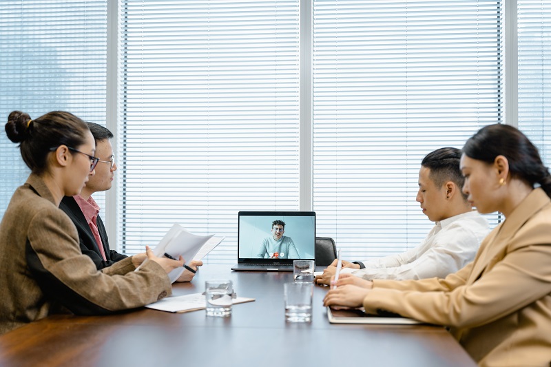 A work meeting with four people sitting physically together in an office and another person joining remotely via a computer.