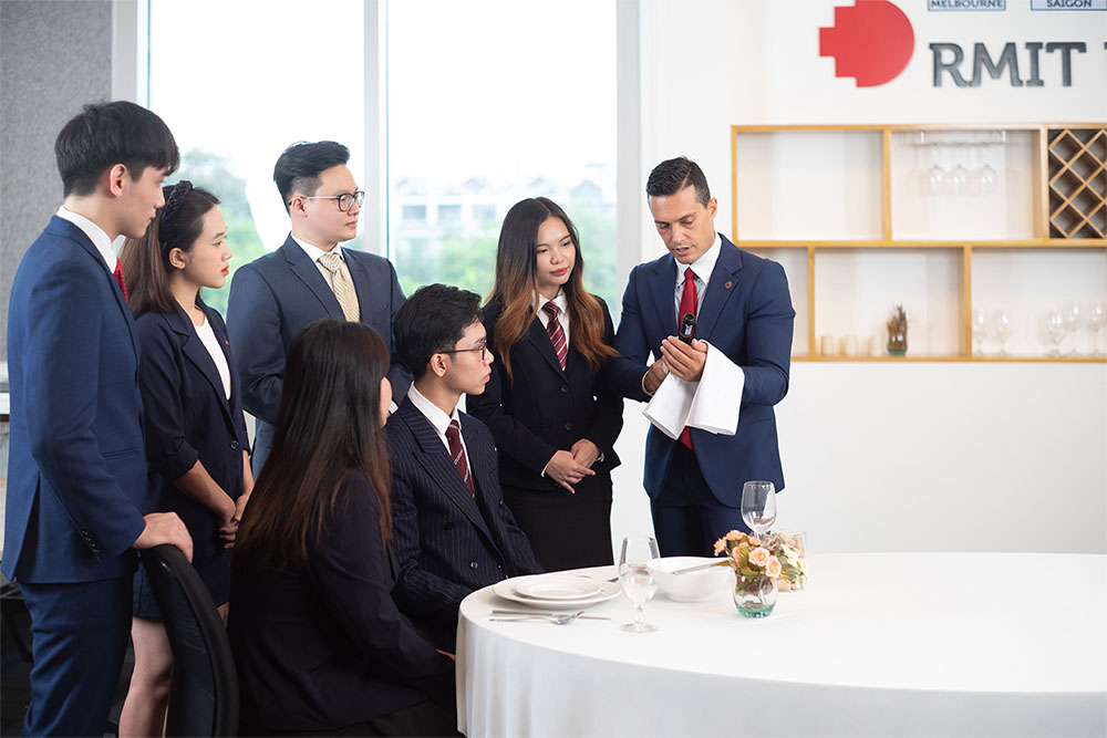 a group of students wearing suits gathering around a round table and listening to a lecturer holding a bottle of wine 