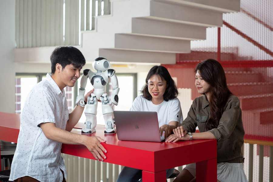 Three RMIT students looking at a laptop beside a robot on a red table