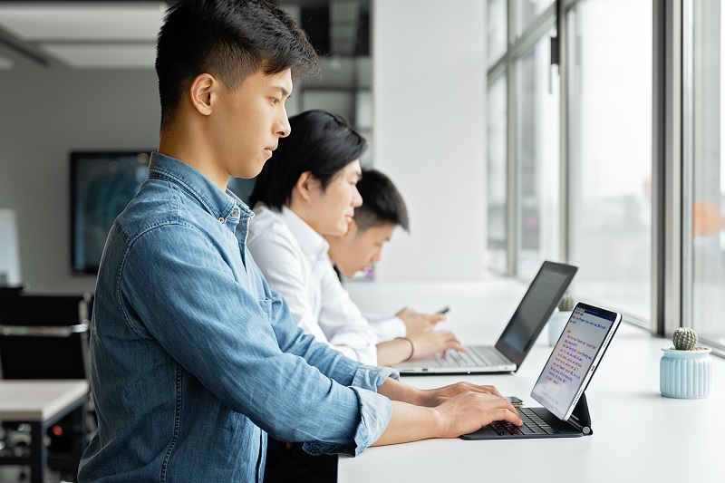 three men working at computers at a window-side long table