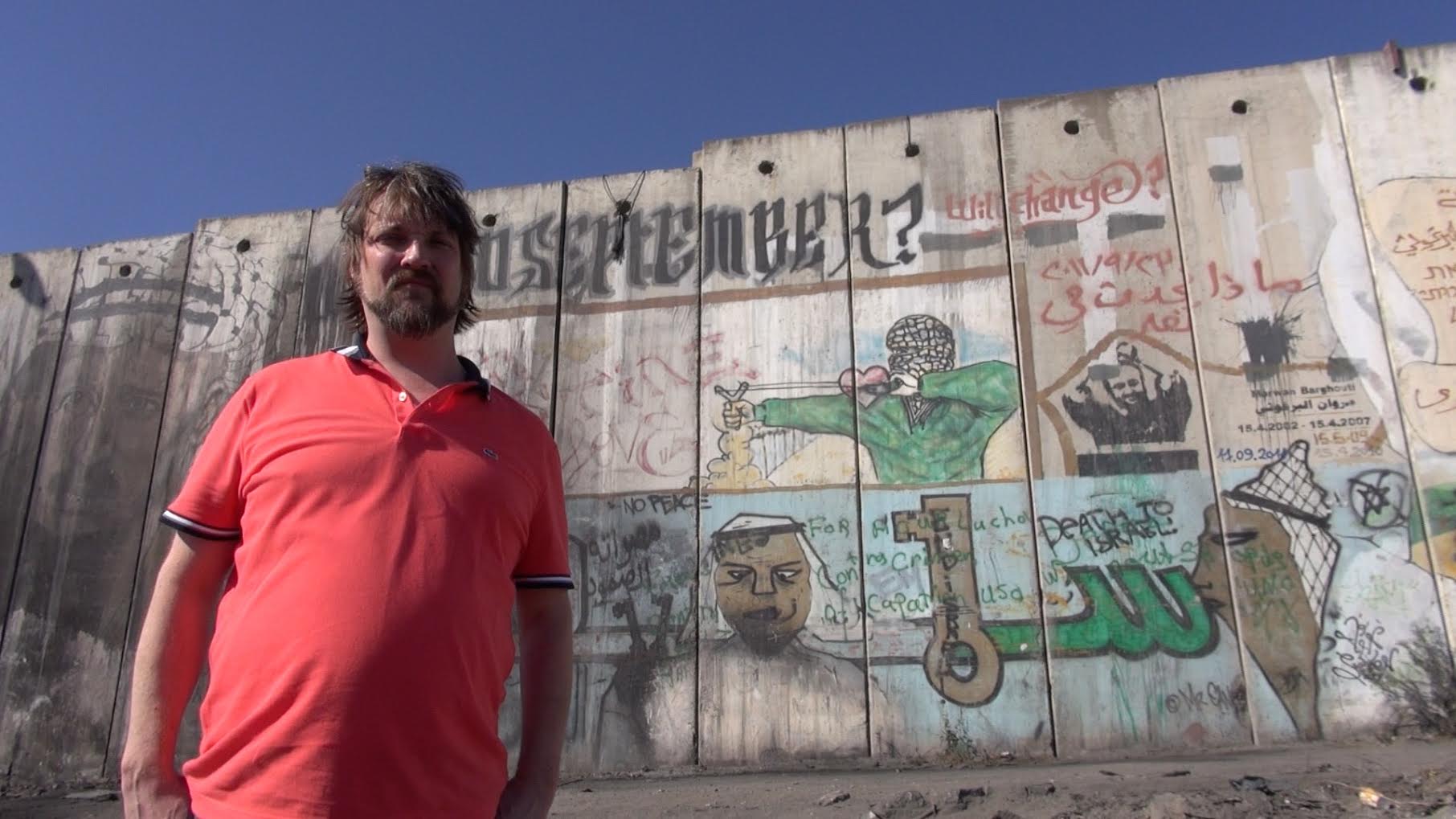 Tiến sĩ Bryan Urbsaitis đứng trước bức tường apartheid chia cắt Palestine và Israel.