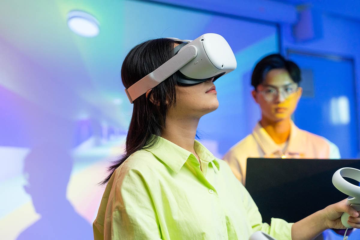Female student in a yellow shirt with hair worn loose and wearing a VR headset  with handsets, in a classroom setting while a male student looks on. 