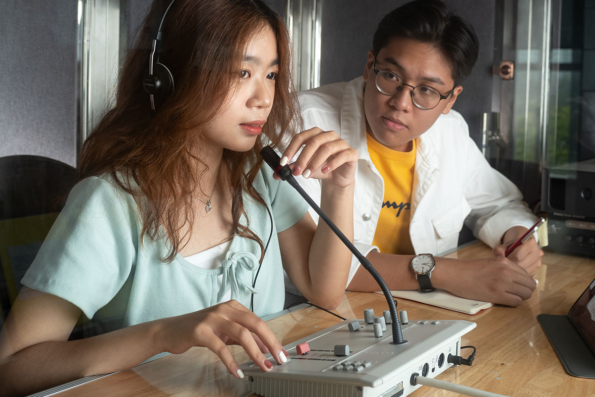 A female student in a green dress does live translation as a male student wearing glasses in a white shirt with a yellow t-shirt underneath takes notes in a notebook