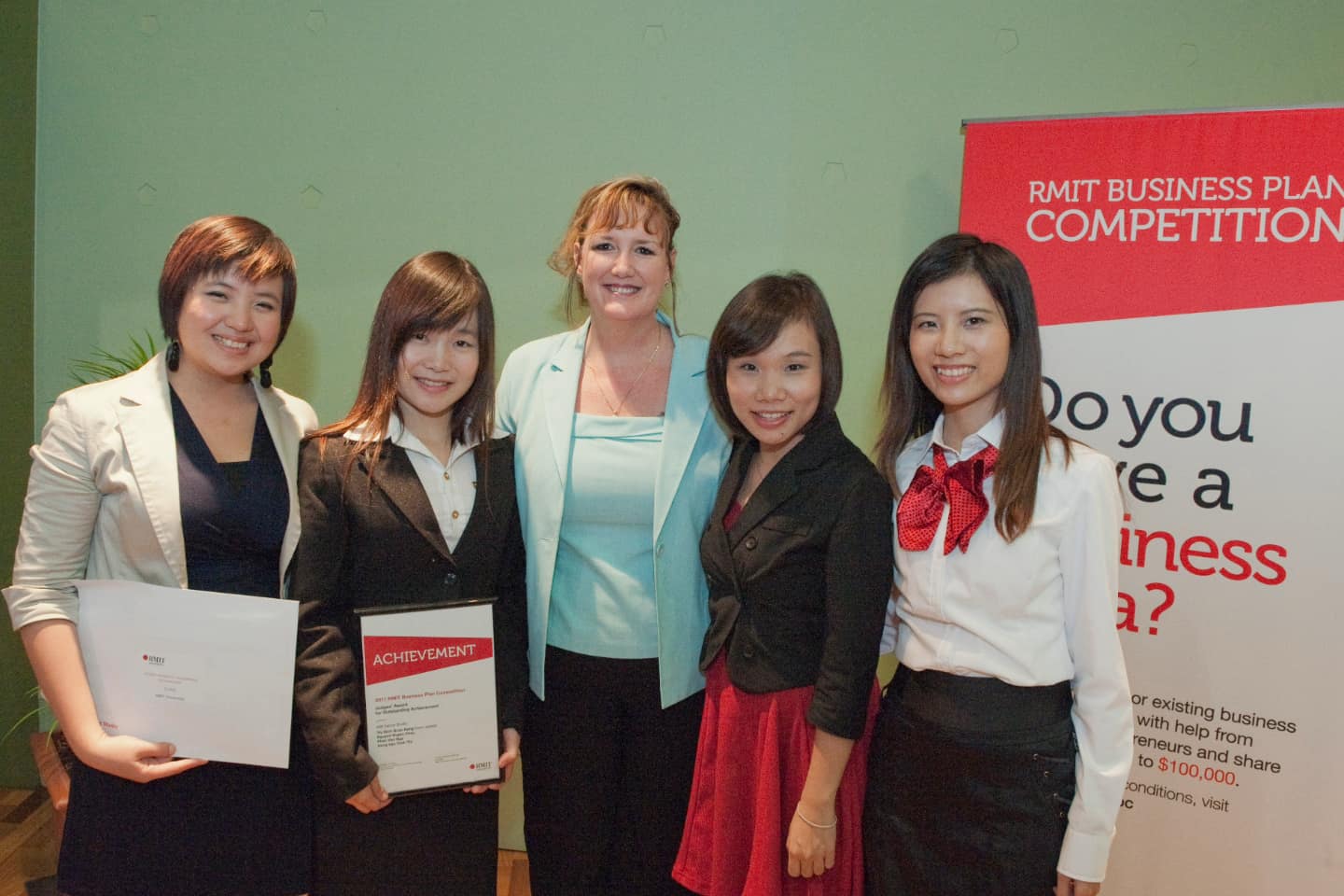 Group of participants at the RMIT Business Plan Competition, holding certificates of achievement, in front of a banner promoting business idea development.