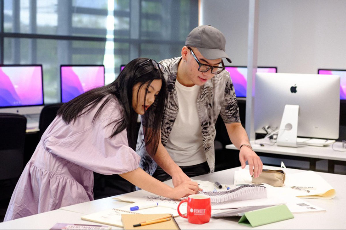 Hai sinh viên cúi xuống bàn trong phòng máy tính, cùng làm việc với tài liệu thiết kế, sổ tay, giấy tờ và ly RMIT màu đỏ