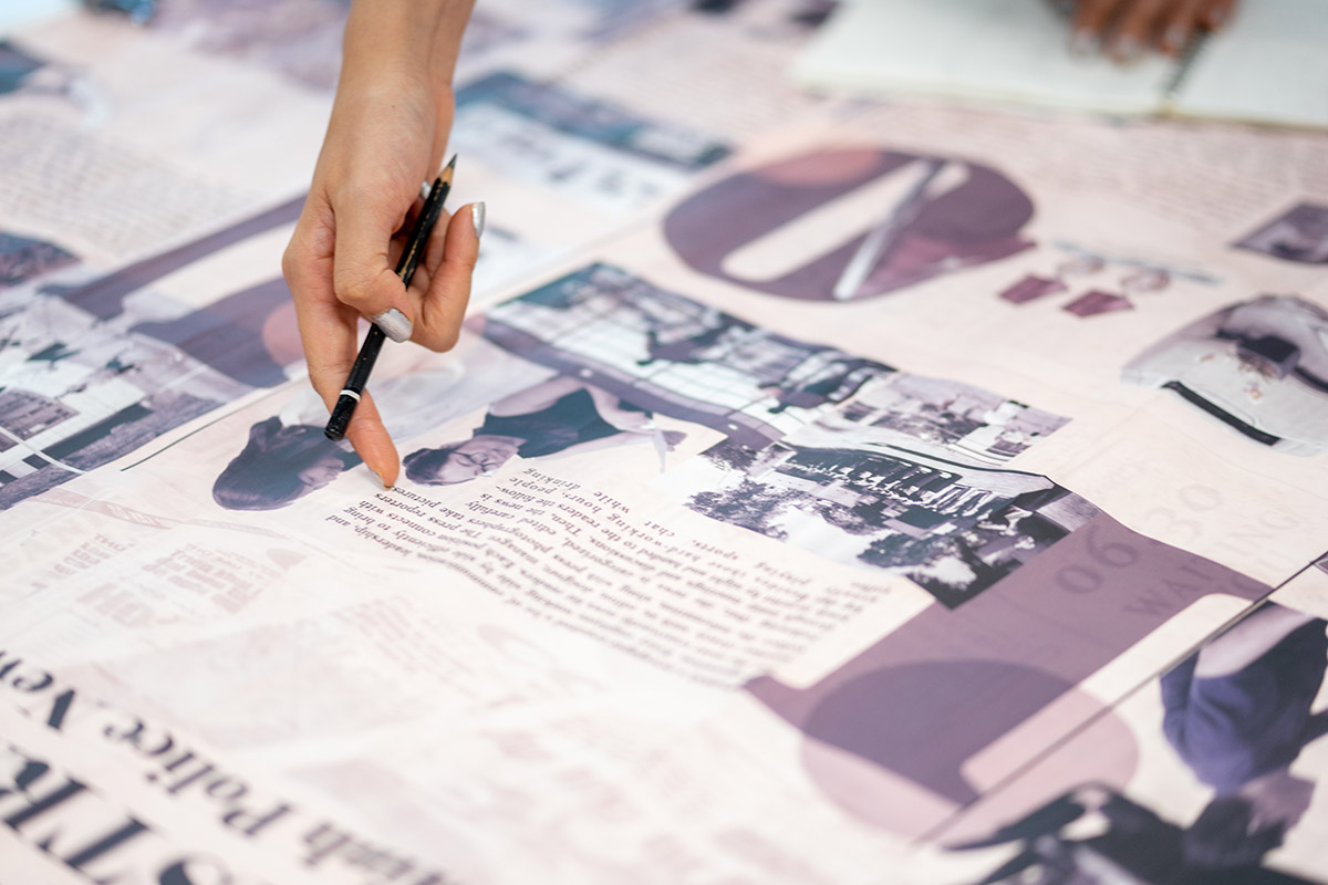 Cận cảnh bàn tay cầm bút chỉ vào bản thiết kế in khổ lớn với hình ảnh và văn bản trải trên bàn