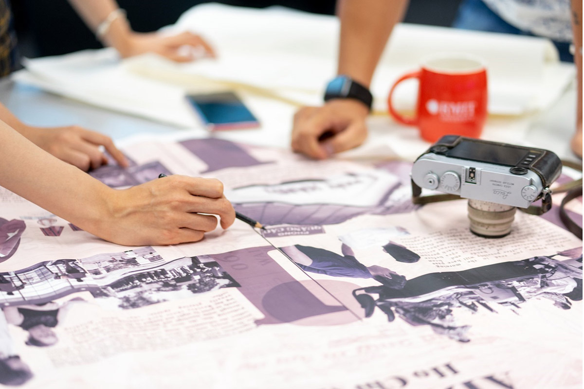 Cận cảnh đôi tay làm việc trên bản thiết kế in khổ lớn với hình ảnh và văn bản trên bàn, bên cạnh có máy ảnh và ly RMIT màu đỏ