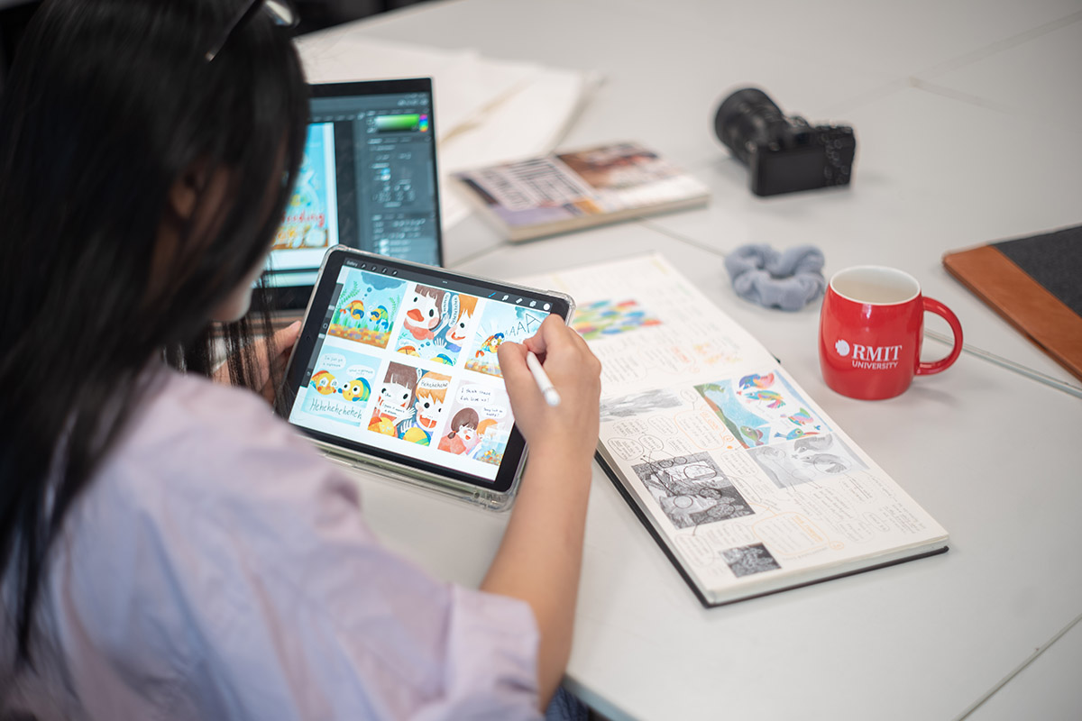 Một sinh viên đang sử dụng máy tính bảng để thiết kế đồ họa màu sắc, trên bàn có sổ phác thảo mở, laptop, máy ảnh và ly RMIT màu đỏ