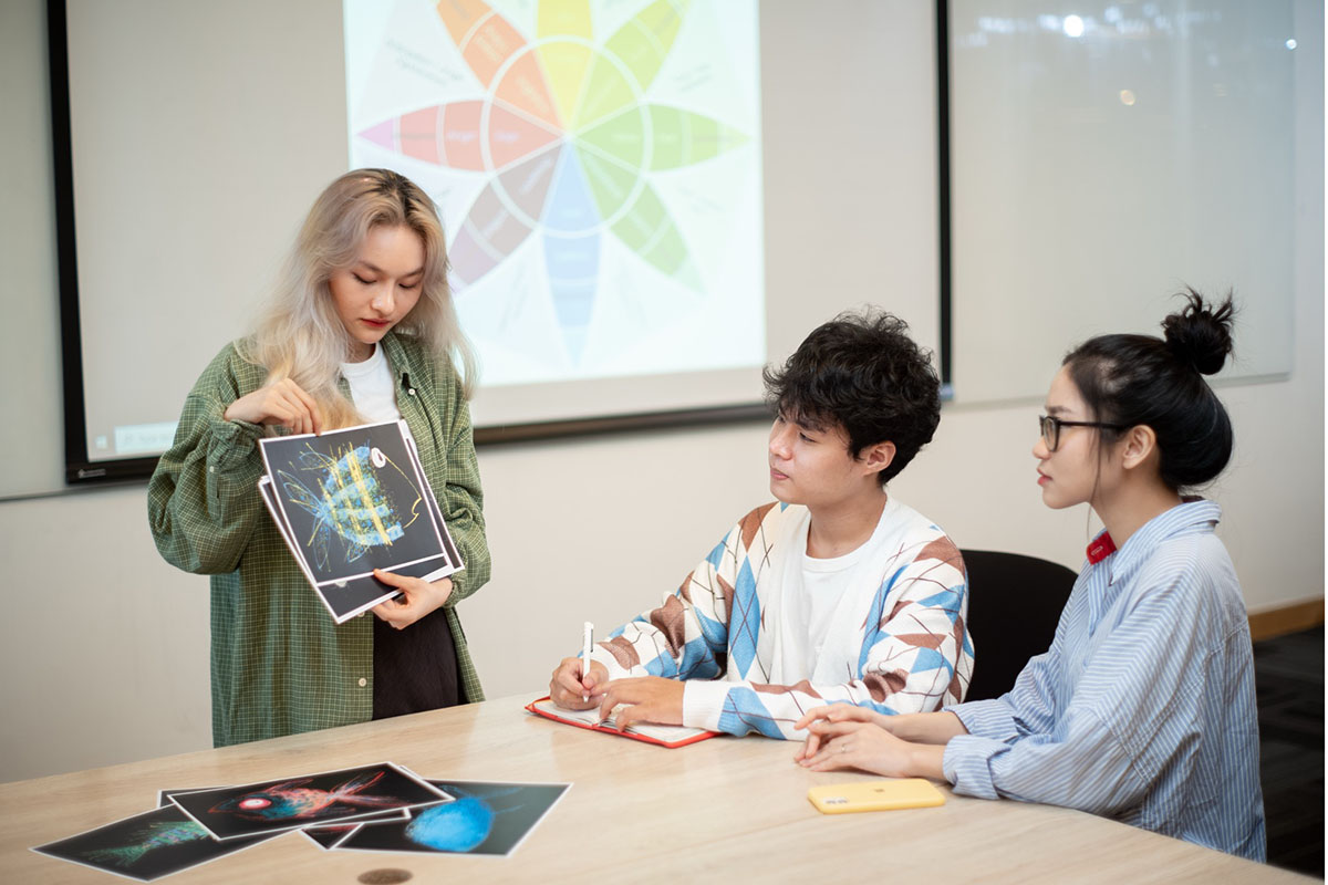 Một sinh viên đang đứng trình bày các hình ảnh in với hai sinh viên khác đang ngồi quanh bàn trong lớp học tâm lý