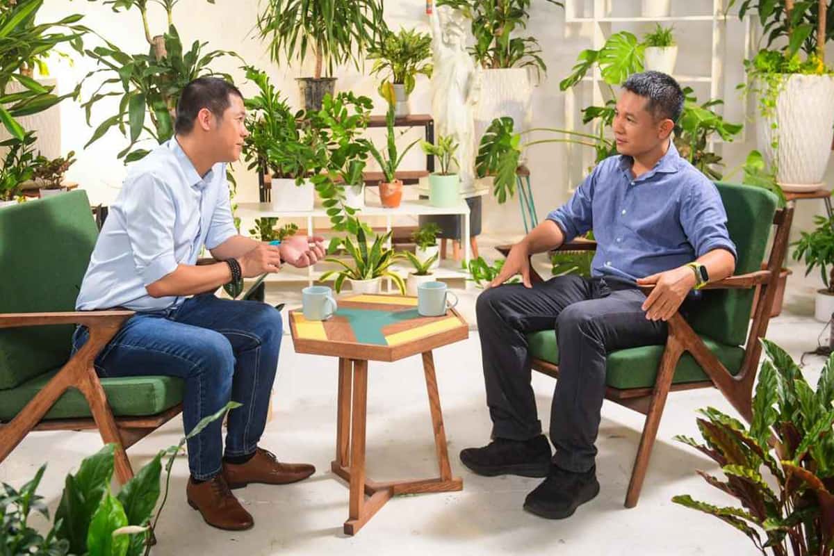 Two Asian men sitting in armchairs and having a conversation in a brightly lit room surrounded by various potted plants, with two coffee mugs on a small table between them.